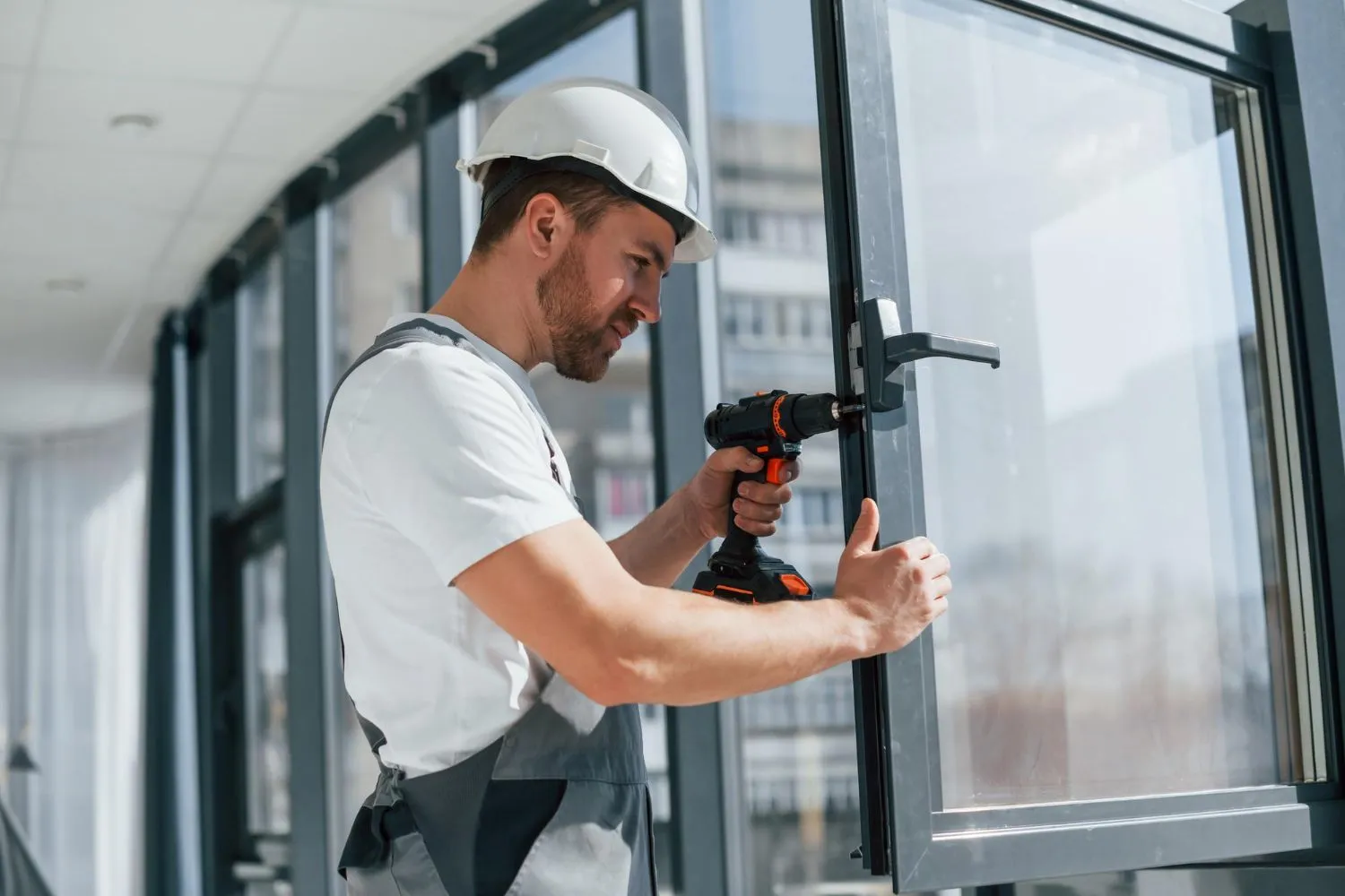 An installer in a hard hat drilling a window frame during glass replacement.